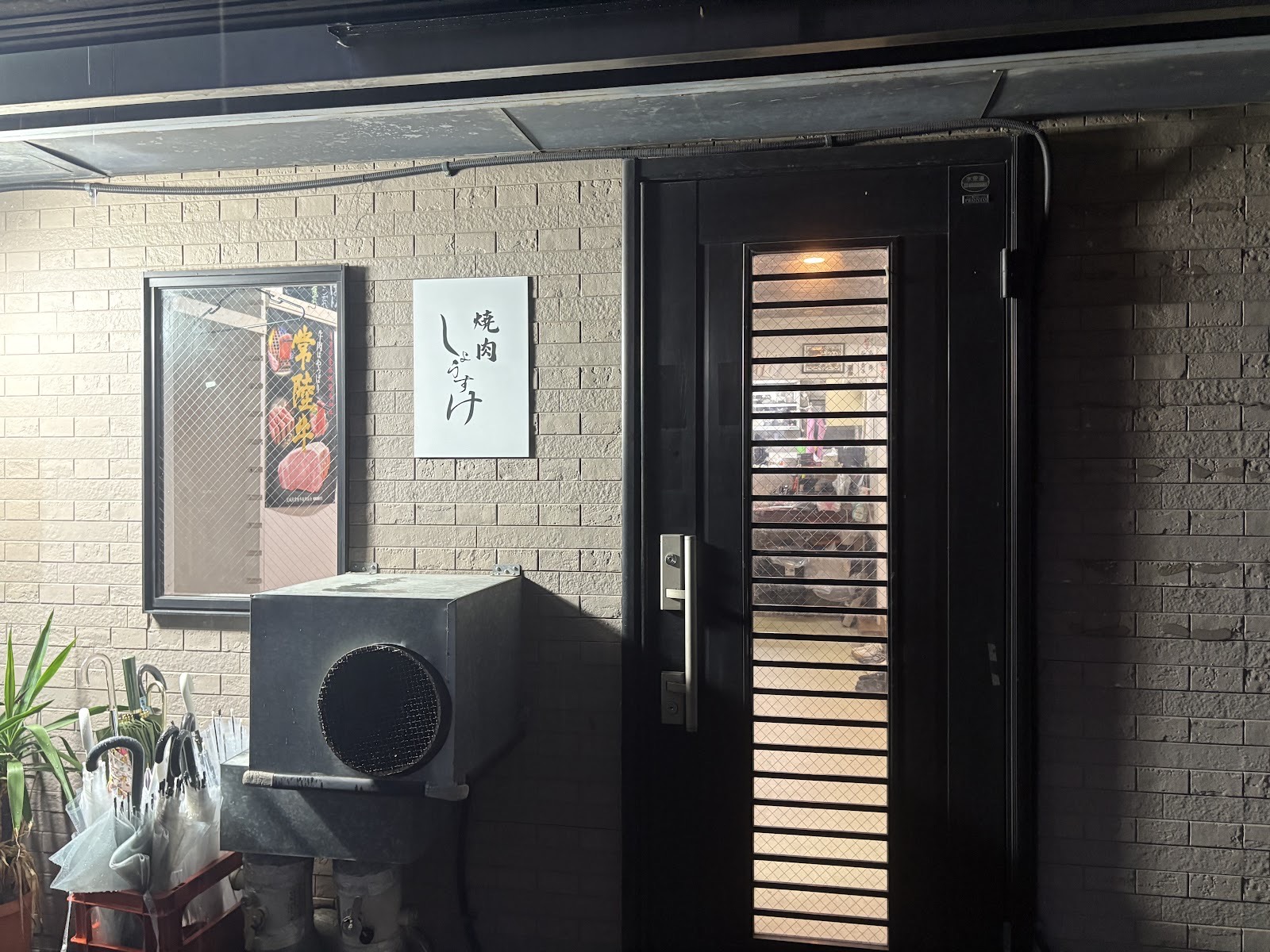 夜の入口。ベージュのタイル壁に「焼肉 しょうすけ」と墨書きした白い看板。黒い扉の格子ガラスから店内の灯りがもれ、左の窓にポスターと傘立て。