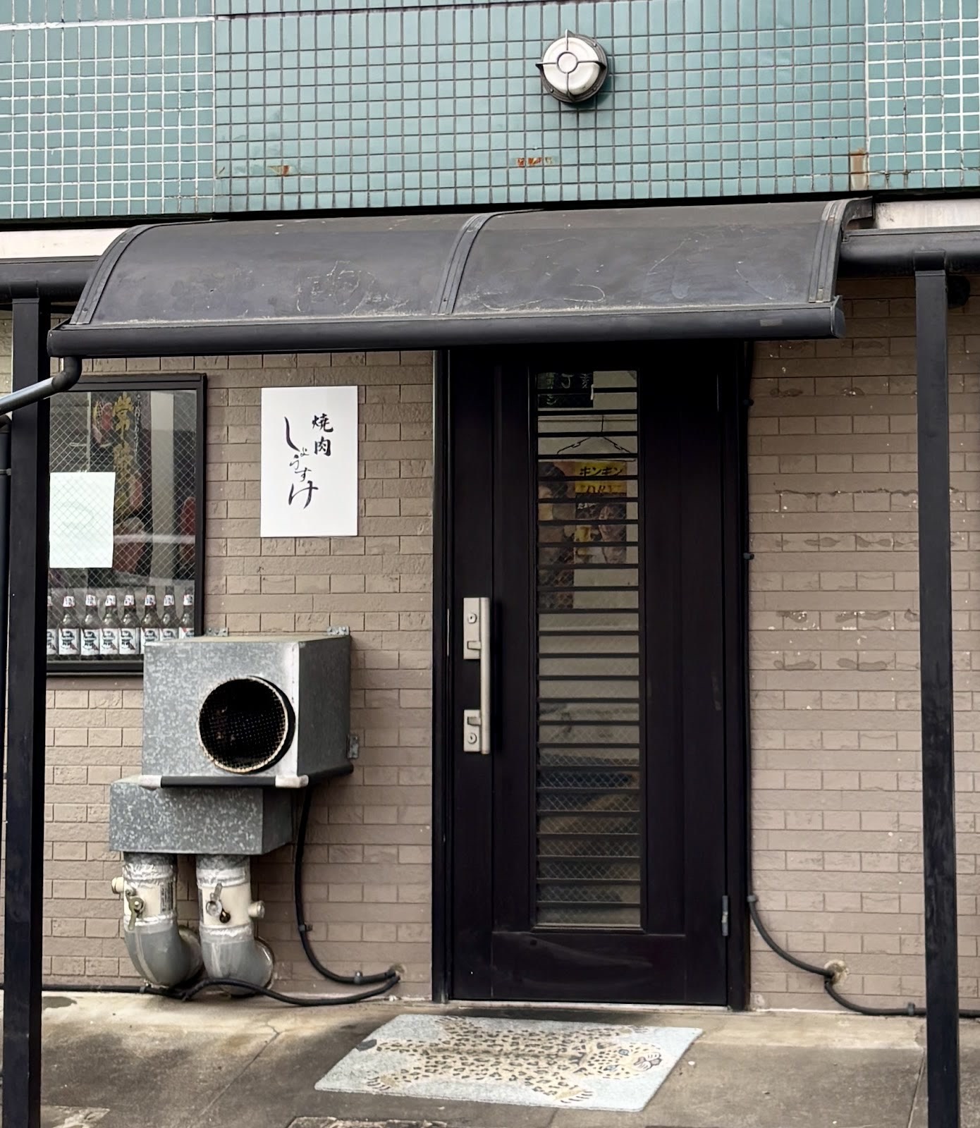 昼の店先。黒い庇の下、黒い格子扉の横に「焼肉 しょうすけ」の白い看板。緑のタイル壁に丸い灯り、足元に豹柄のマット。