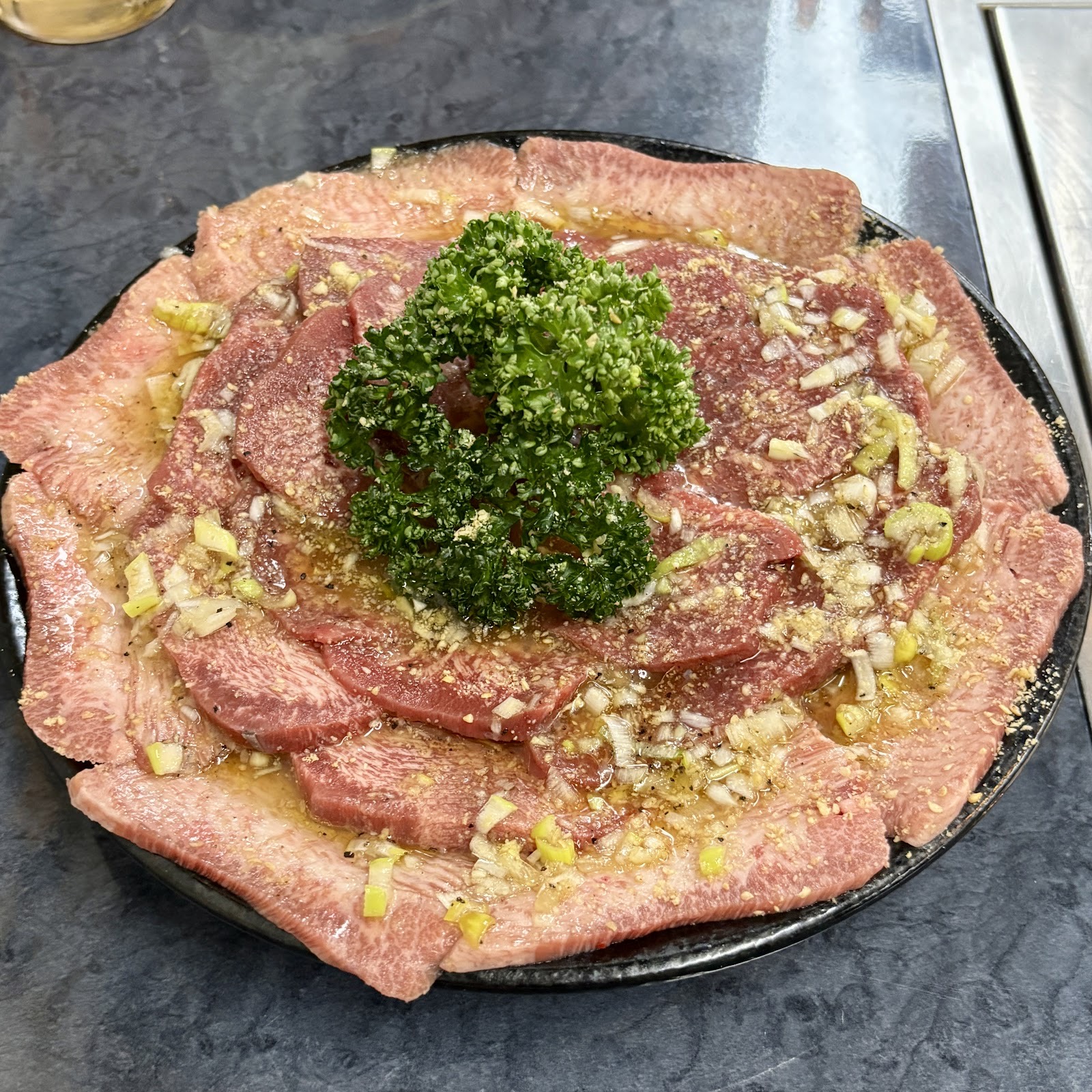 黒い大皿に薄切りの生肉を円く並べ、刻みねぎと油だれ、中央にパセリ。青い石目のテーブル。