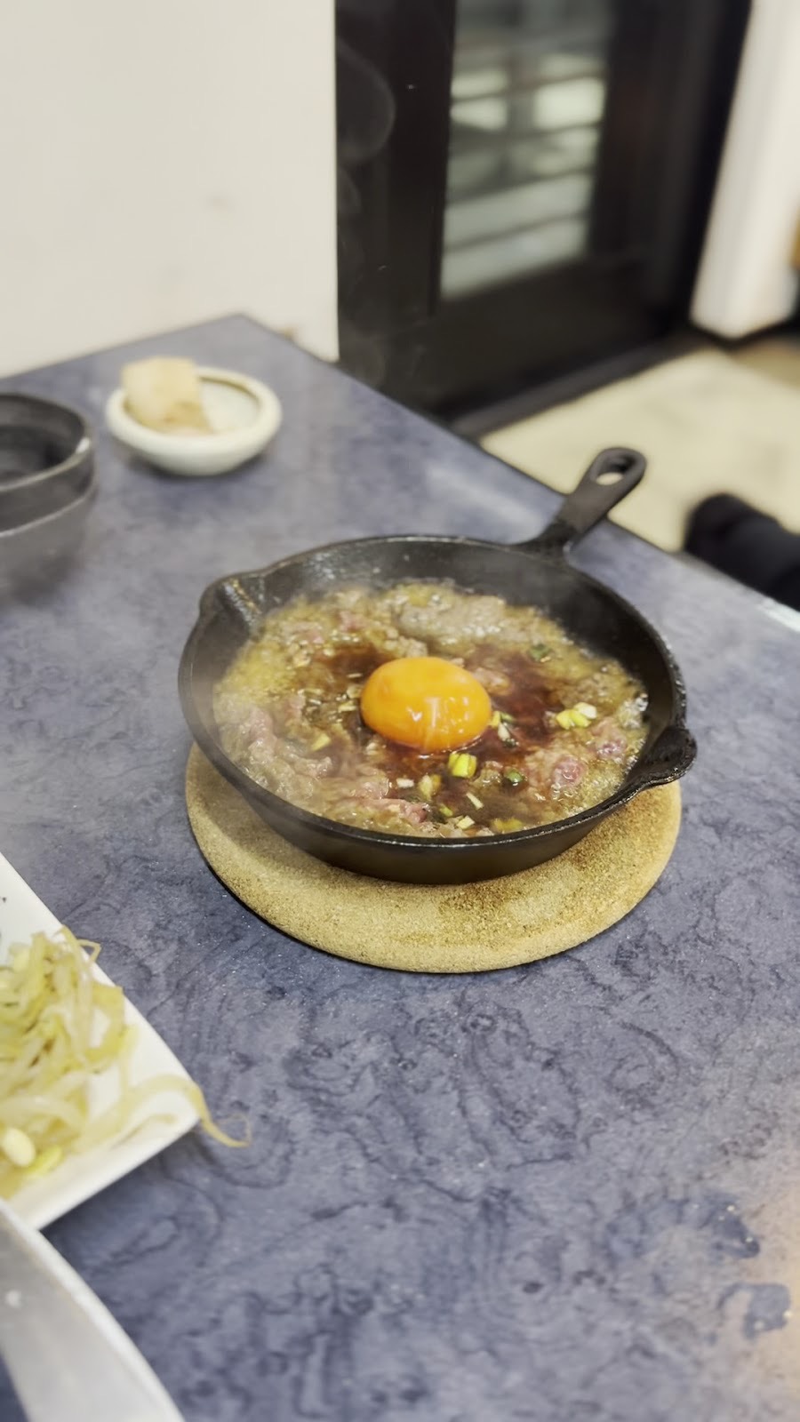 青い石目のテーブルに、コルクの敷き板にのせた小さな鉄のスキレット。たれの肉に卵黄、左にもやしの小皿、奥に格子ガラスの扉。