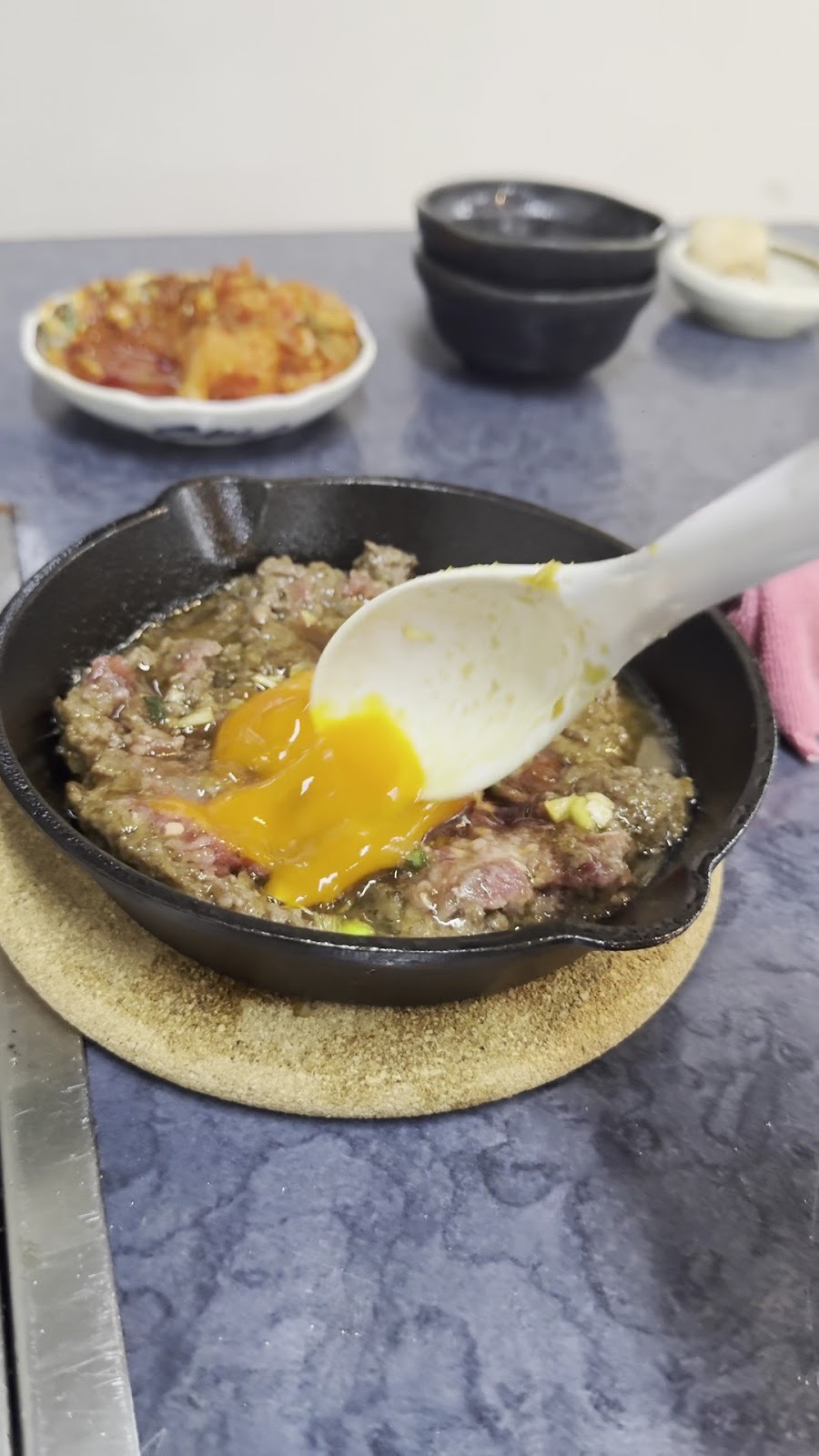 鉄のスキレットの中で、白いスプーンが卵黄をたれの肉に混ぜている。奥に赤い和え物の小鉢と重ねた黒い器。