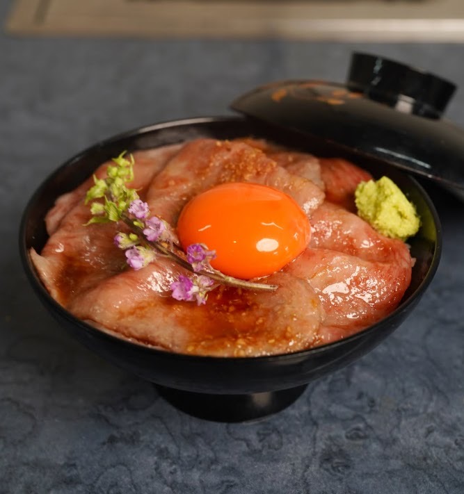 黒い漆の丼に薄切りの生肉を敷き、中央に卵黄、わさびと小さな花。横に蓋。